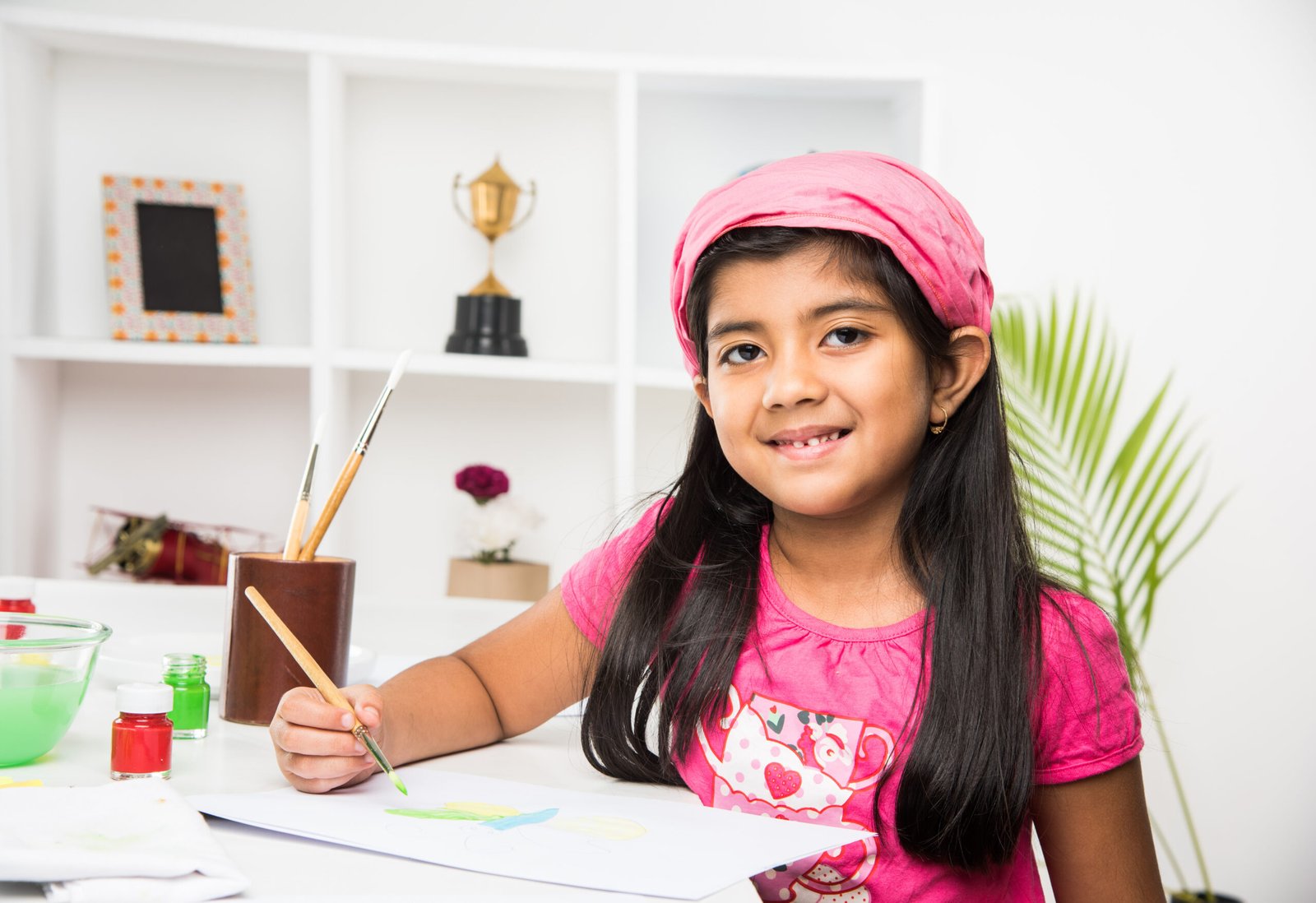 Cute Little Indian or Asian girl child enjoying drawing OR painting with brush and paint over paper at home