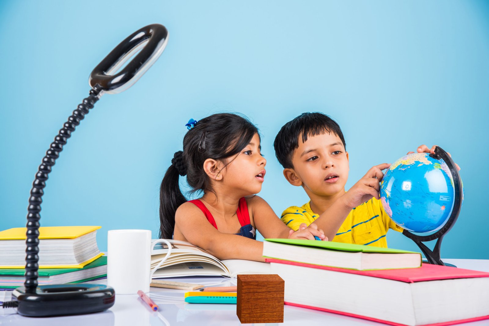 Cute little Indian or Asian kids studying on study table with pile of books, educational globe, isolated over light blue colour